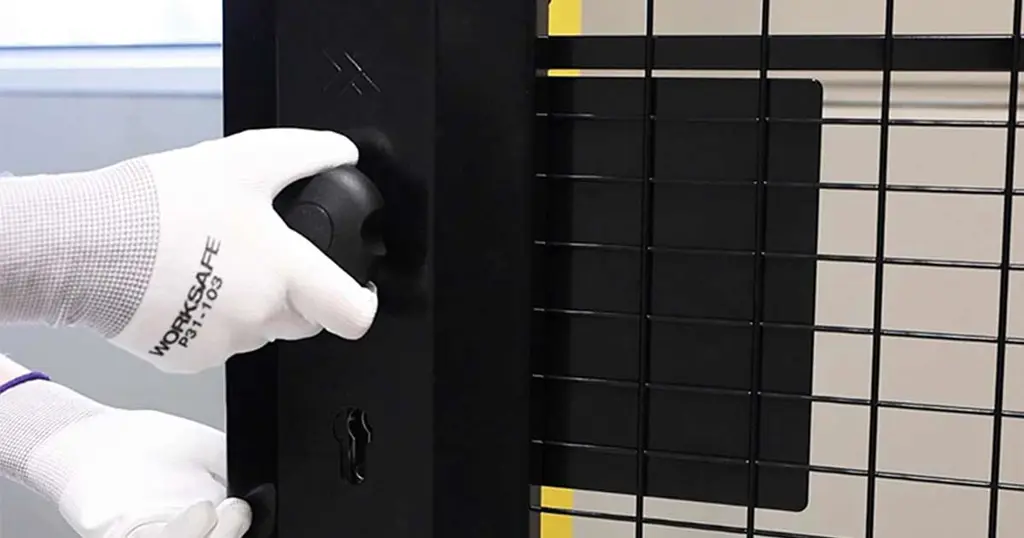 Mano sosteniendo una cerradura de cilindro con apertura de emergencia montada en un panel de malla negro 